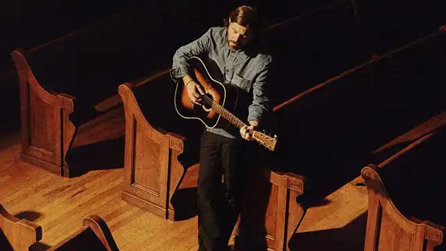 Josh Baldwin playing acoustic guitar alone among wooden church pews